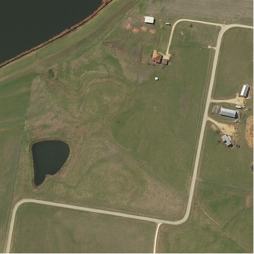 Aerial view of the property: open pasture with a pond at the southwest corner, a house and outbuildings near the north edge, and county roads along the east and south sides.